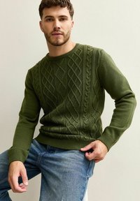 Zielony sweter dzianinowy z wzorem w warkocze, okrągłym dekoltem i ściągaczami. W zestawieniu z niebieskimi dżinsami.