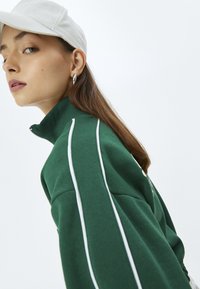 Sudadera verde con cremallera, con rayas blancas a lo largo de las mangas, tejido texturizado, cuello alto y detalle bordado, combinada con una gorra blanca.