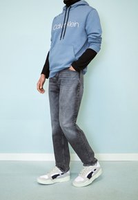 Ljusblå hoodie med vitt logotyp, bärs över en svart långärmad tröja. Gråjeans och vita sneakers med svarta accenter fulländar looken.