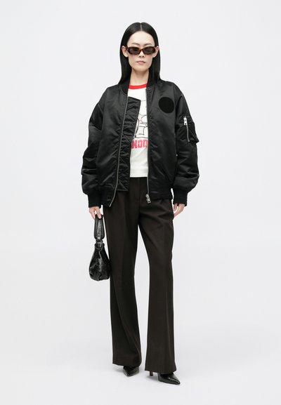 Crna bomber jakna s okruglim izvezom, bijela majica s grafikom i crvenim detaljima, smeđe hlače visokog struka i crne špicaste štikle.