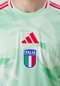Žalias sportinis džemperis su subtiliais baltais raštais, raudonas Adidas logotipas ir Italijos nacionalinės komandos ženkliukas, kuriame dominuoja mėlyna, balta ir raudona spalvos.