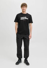Sort T-shirt med hvid "JACK & JONES" tekst, parret med løse sorte bukser og sorte sneakers med blå detaljer. Enkel, afslappet outfit.