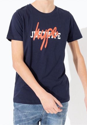 Persoon die een marineblauw T-shirt draagt met witte en oranje tekst "JUPE" en "hype" die elkaar overlappen, gecombineerd met lichtblauwe jeans.