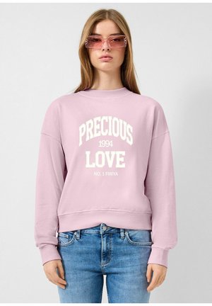 Kobieta w różowej bluzie z napisem "PRECIOUS 1994 LOVE NO. 1 FIWYA", niebieskich dżinsach oraz różowych okularach przeciwsłonecznych w prostokątnym kształcie, stojąca na białym tle.