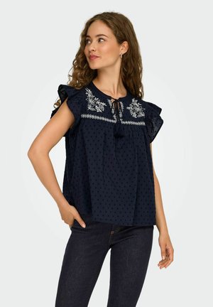 Blouse bleu marine avec des manches courtes à volants, texture à pois et broderie florale blanche au niveau du col. Présente un détail de lien à l'avant.