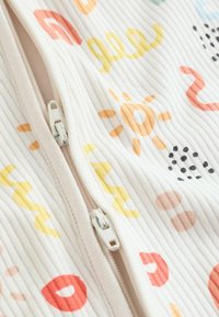 Geripptes Stoffmaterial mit mehrfarbigen abstrakten Mustern, darunter Sonnen, Wirbel und Punkte; zentraler weißer Reißverschluss mit zwei Zippern.