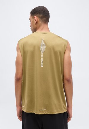 Hátraforduló férfi, aki ujjatlan olívazöld pólót visel, amelyen függőleges „NEVER STOP RUNNING” felirat és cipőnyom grafikája található a hátoldalon.