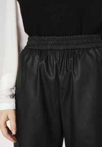 Pantalones de imitación de cuero negro, con pliegues y cinturilla elástica, combinados con una blusa blanca de manga larga. Textura suave con un acabado brillante.
