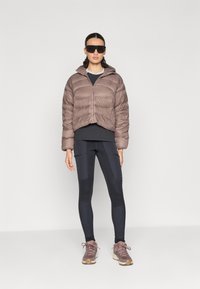 Brun kort puffjacka med huva, ribbad mudd, bärs över en mörk topp och svarta leggings. Matt svarta solglasögon och gråbruna sneakers.