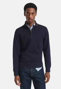 Sudadera de lana en color azul marino hecha de algodón, con un cuello de media cremallera y un acento marrón. Presenta puños y dobladillo acanalados, llevada sobre una camisa de cuadros azul.