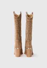 Une paire de bottes de cowboy en daim beige avec des broderies décoratives, des clous en métal et des talons en bois empilés, montrées de l'arrière.