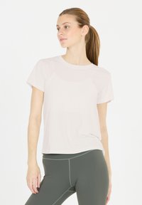 Mujer joven con el cabello en una coleta, vistiendo una camiseta de manga corta rosa claro y leggings grises oscuros, de pie contra un fondo blanco.