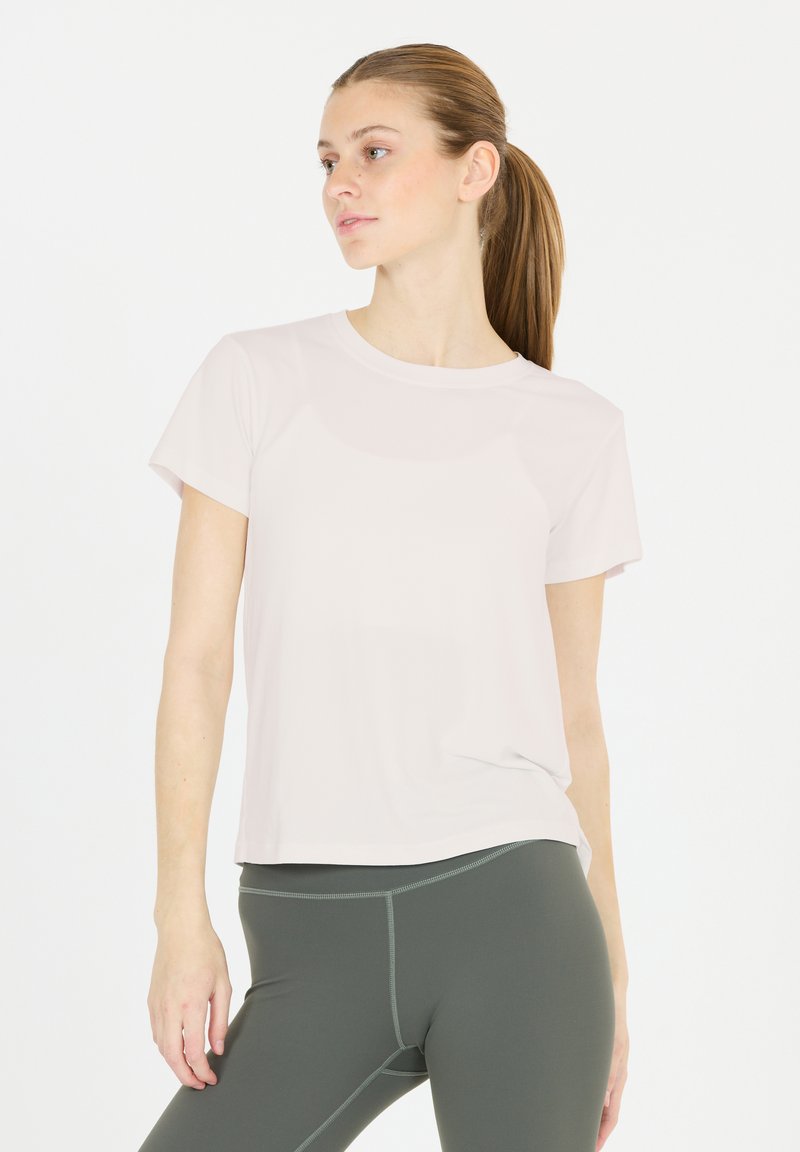 Mujer joven con el cabello en una coleta, vistiendo una camiseta de manga corta rosa claro y leggings grises oscuros, de pie contra un fondo blanco.