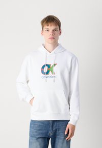 Hoodie blanc avec une poche avant, cordon de serrage, et un logo "OK" coloré aux accents bleu et vert, associé à un jean bleu.
