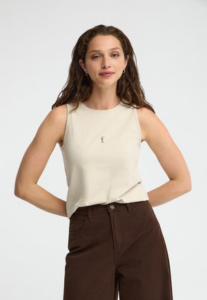 Joven mujer con cabello largo y rizado que lleva una camiseta sin mangas beige y pantalones marrones de cintura alta sobre un fondo blanco liso.