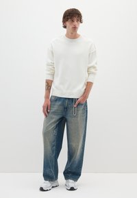 Witte gebreide trui met geribde manchetten en zoom, gecombineerd met blauwe baggy jeans met een vervaagde wash en kettingdetail. Schoeisel zijn witte sneakers.