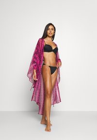 Rožnat prosojni kimono s teksturiranim dizajnom, v kombinaciji s črnim bikini. Kimono ima ohlapen kroj in se ob robu telesa prosto spušča.