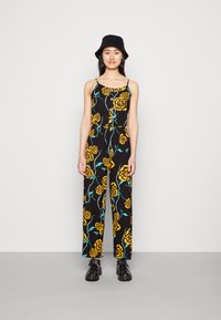 Macacão floral com fundo preto, apresentando flores amarelas e caules azul-turquesa. Design com alças, corpo ajustado e calças largas. Sapatos pretos.