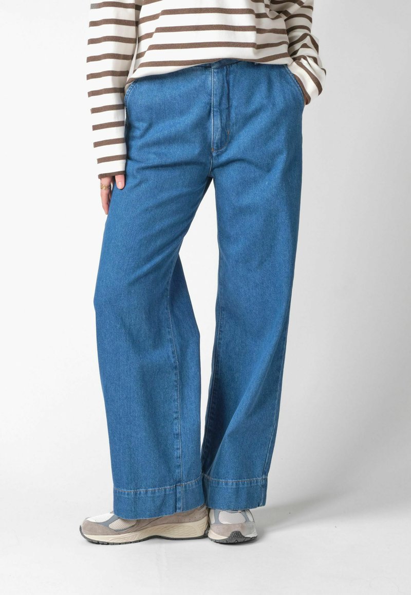 Persona que lleva pantalones vaqueros anchos azules, zapatillas beige y blancas, y una camiseta de manga larga con rayas beige y marrones sobre un fondo liso.