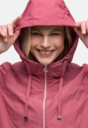 Personne souriante portant une veste à capuche rose zippée, tenant les bords de la capuche avec les deux mains sur un fond clair uni.