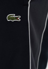 Κεντητό πράσινο λογότυπο κροκοδείλου με κόκκινο στόμα σε μαύρο ύφασμα με κάθετη λευκή επένδυση.