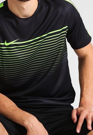 Mies, jolla on päällään musta Nike-urheilupaita, jossa on neonvihreitä raitoja ja logo, istuu kädet polvilla.