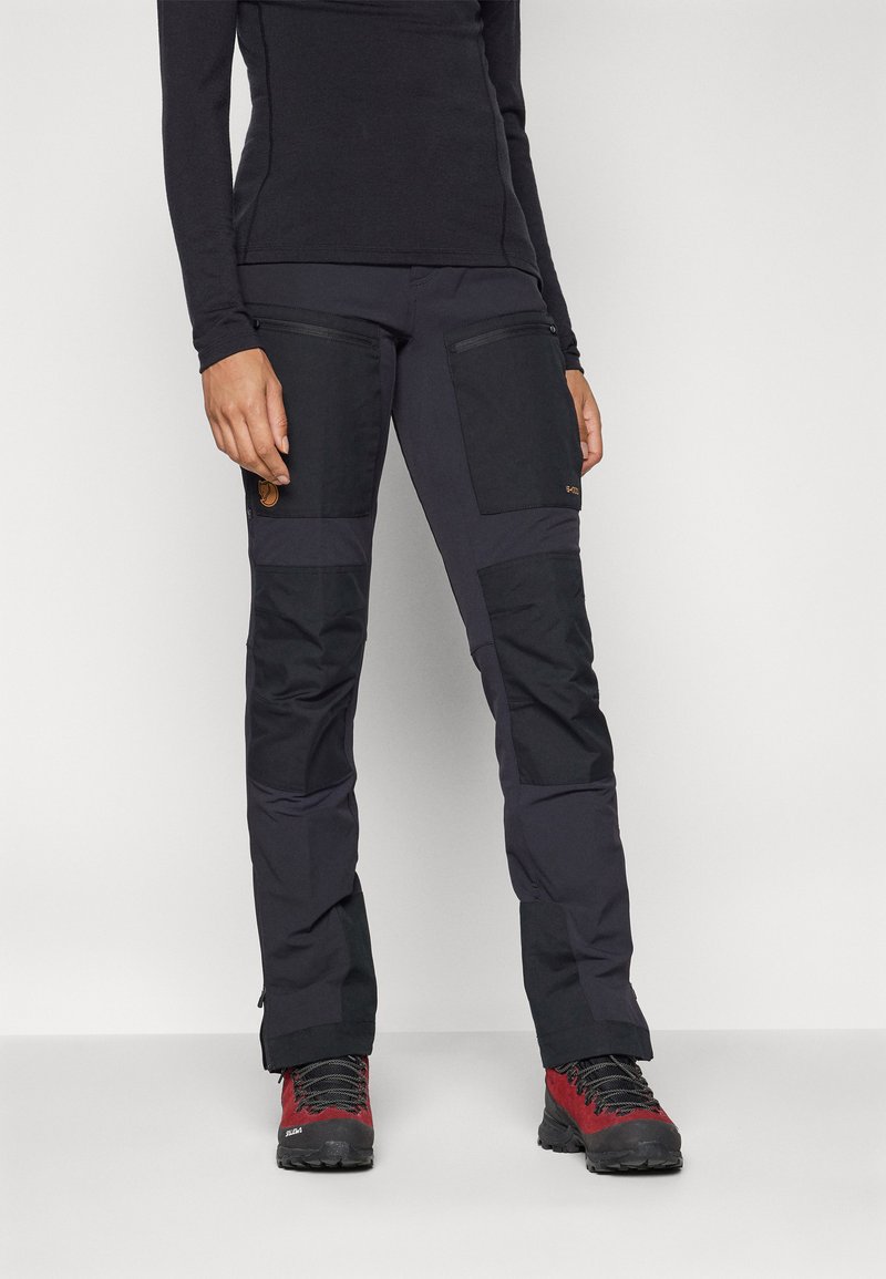 Zwarte softshell broek met ritszakken, versterkte knieën en een getailleerd ontwerp, gecombineerd met rode en zwarte wandelschoenen.