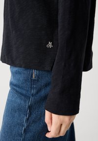 Person wearing dark long-sleeve top with small "M O'P" logo near hem and blue denim jeans, hand relaxed by side against a neutral background.