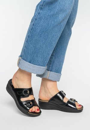 Black patent leather sandals with dual circular buckle accents and a wedge sole, paired with cuffed blue denim jeans.