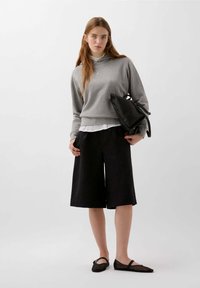 Felpa grigia con un design cropped, indossata sopra una maglietta bianca. Pantaloni shorts larghi neri e scarpe flat nere completano l'outfit. Stile minimalista.