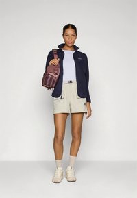 Kurtka polarowa w kolorze ciemnogranatowym, zestawiona z beżowymi szortami. Akcesoria obejmują bordową torbę na ramię. Modelka stoi z neutralnym wyrazem twarzy.