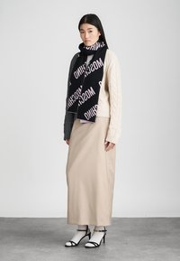 Suéter de malha em creme com padrão de tranças, combinado com uma saia maxi bege. Cachecol preto com "MOSCHINO" em rosa. Saltos altos pretos com tiras.