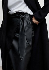 Jupe en cuir noir avec un détail de nœud noué à la taille, texture lisse, portée avec un haut blanc et un manteau noir.