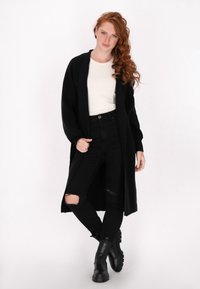 Zwarte lange cardigan van textuur materiaal, met een open voorkant, gecombineerd met high-waisted zwarte jeans met scheuren en zwarte enkellaarsjes.