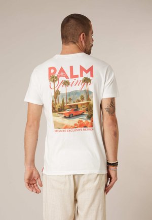 Man wearing white t-shirt featuring a Palm Springs retro car and palm tree graphic with beige pants and arm bracelets.