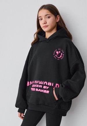 Ragazza adolescente che indossa una felpa nera oversize con faccina rosa sorridente e la scritta "Baker by Ted Baker", in piedi contro una parete chiara e semplice.