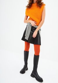 Mujer vestida con un suéter sin mangas naranja, una falda negra con pliegues, medias naranja, botas negras hasta la rodilla y una camisa a rayas atada a la cintura.