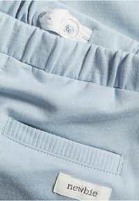 Lichtblauwe stof met een geplooide elastische tailleband, voorzien van een witte knoopdetail en een geborduurd "newbie" label. Gladde textuur, casual ontwerp.