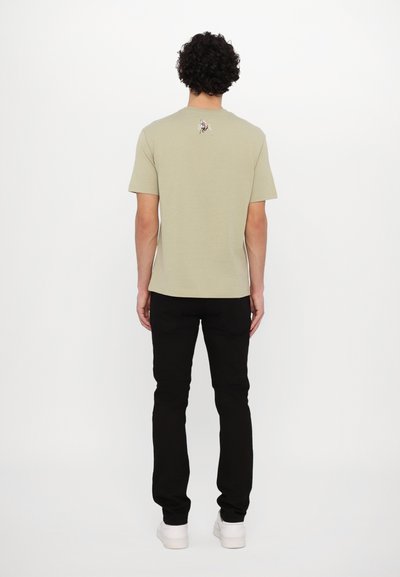 T-shirt en coton vert olive avec un col rond et des manches courtes, arborant un petit motif brodé sur le haut du dos. Porté avec un pantalon noir.