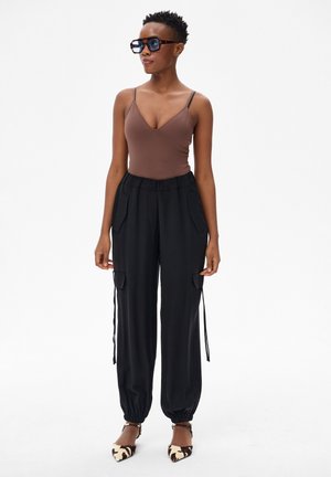 Kobieta w okularach przeciwsłonecznych w oprawkach w panterkę, brązowej bluzce bez rękawów, czarnych spodniach cargo ze ściągaczami i butach w zwierzęcy wzór ze spiczastymi czubkami stojąca na tle białego tła.