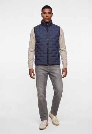 Mann trägt eine marineblaue gesteppte Weste über einem beigen Pullover mit grauen Jeans und beigen Sneakers und steht vor einem weißen Hintergrund.