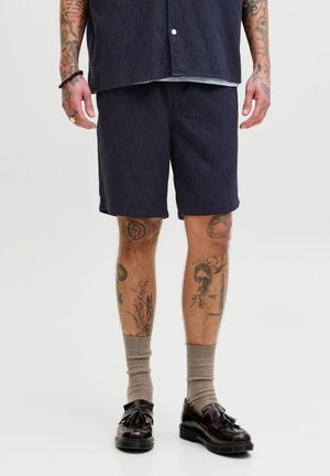 Homme portant un short texturé foncé, des chaussettes beiges et des mocassins à glands foncés, avec des jambes et des bras tatoués visibles sur un fond uni.