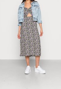 Mustat kukkakuosiset midimekot, joissa on pieniä purppuran ja valkoisen värisiä kukkia, rinnassa solmu yksityiskohta, yhdistettynä vaaleaan farkkutakkiin ja valkoisiin lenkkareihin.