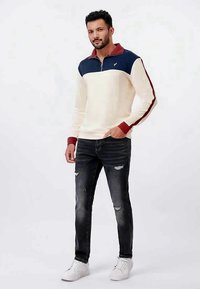 Crème zip-up pullover met marineblauwe en bordeauxrode accenten, gecombineerd met versleten donkere jeans en witte sneakers. Gladde textuur en casual pasvorm.