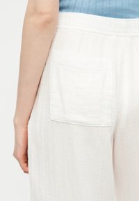 Κοντινό πλάνο ενός ατόμου που φοράει λευκό υφασμάτινο παντελόνι με πίσω τσέπη και ανοιχτό μπλε πλεκτό τοπ, δείχνοντας το αριστερό χέρι και γοφό.