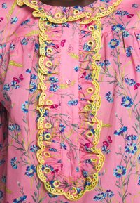 Rosa blus med blommönster och gult broderat kant. Har blå och lila blommor med fjärilar, knäppning fram och volangkrage.