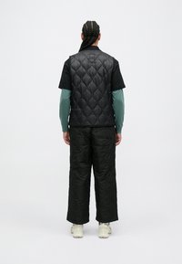 Chaleco acolchado negro sobre una camiseta negra, combinado con pantalones oscuros de corte amplio y zapatillas de color claro, visto desde atrás.