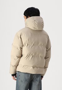 Beige pufferjack met een hoge capuchon, gequiltde textuur en geribbelde manchetten, gecombineerd met blauwe denim jeans. Vanachter gefotografeerd tegen een lichte achtergrond.
