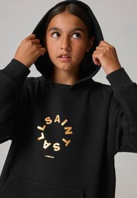 Sudadera negra con bolsillo frontal, con una impresión circular dorada que dice "SAINT LONDON". Tejido texturizado y capucha ajustable.