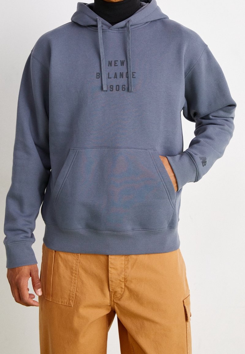 Felpa con cappuccio in morbido tessuto grigio con tasca frontale. Presenta il testo nero "New Balance 906" e polsini e orlo a coste.